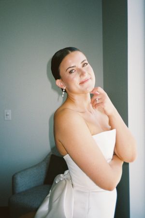 A person in a white strapless dress smiles while getting ready in a room with gray walls.