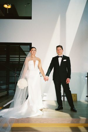 A bride in a white gown with long veil and groom in black tuxedo hold hands in a modern white interior space.