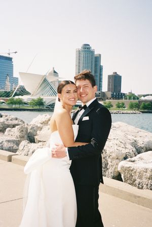 A couple in wedding attire embraces on a waterfront with modern city buildings and architecture in the background.