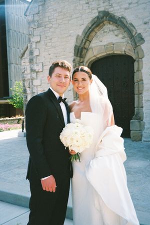 A bride and groom pose together in front of a stone church wall with arched doorways on their wedding day.