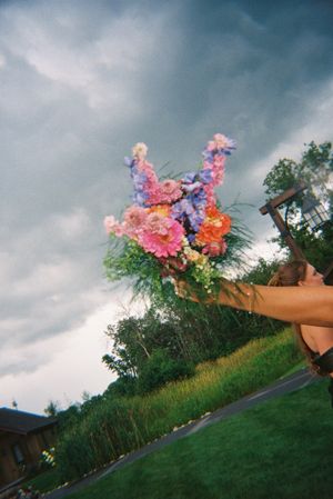 A vibrant bouquet of pink, purple, and orange flowers held up against a dramatic cloudy sky.