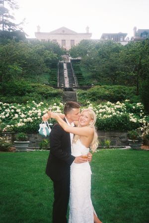 A couple dances together in a garden setting with white flowers and stone walls in the background.