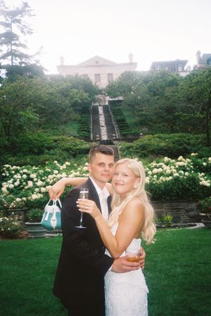 A couple celebrates with drinks in formal attire at a garden venue with grand stairs and white flowers in the background.