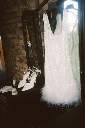 A white sequined cocktail dress with feather trim hem hangs next to matching white high heel shoes against a brick wall.