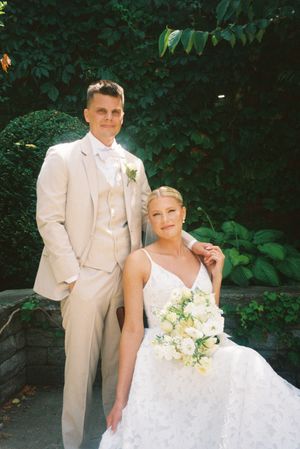 A couple in white wedding attire pose together outdoors with lush greenery in the background.