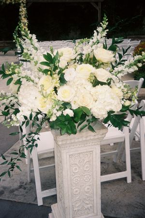 White floral arrangements in ornate carved pedestals create an elegant wedding ceremony display.