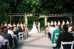 Outdoor garden wedding ceremony setup with white chairs arranged in rows beneath tall trees and natural greenery.
