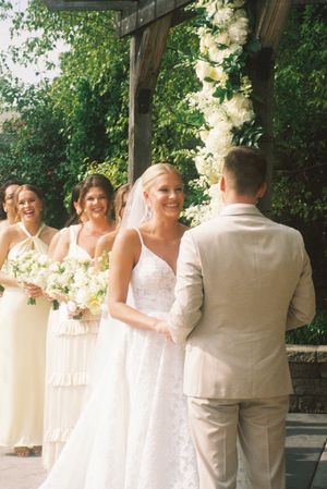 Wedding party in white and cream attire poses outdoors under a covered walkway.