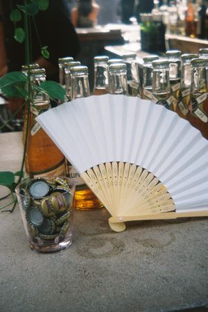 A white paper hand fan with wooden ribs shown from multiple angles on a textured surface next to glassware.