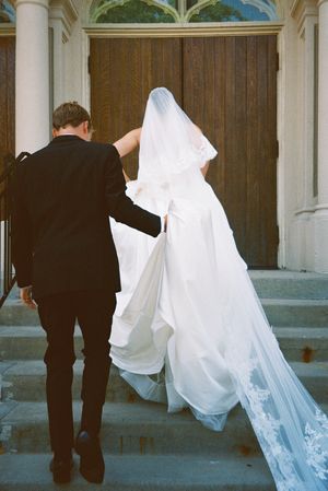A couple enters a church doorway, with the bride's white wedding gown and veil flowing behind them.