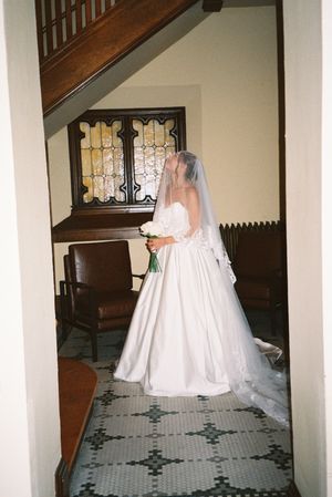 A flowing white wedding dress moves through an elegant hallway with tiled floors and wooden banisters.