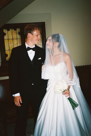 Wedding couple poses together in a church hallway with the bride holding a bouquet and wearing a flowing veil.