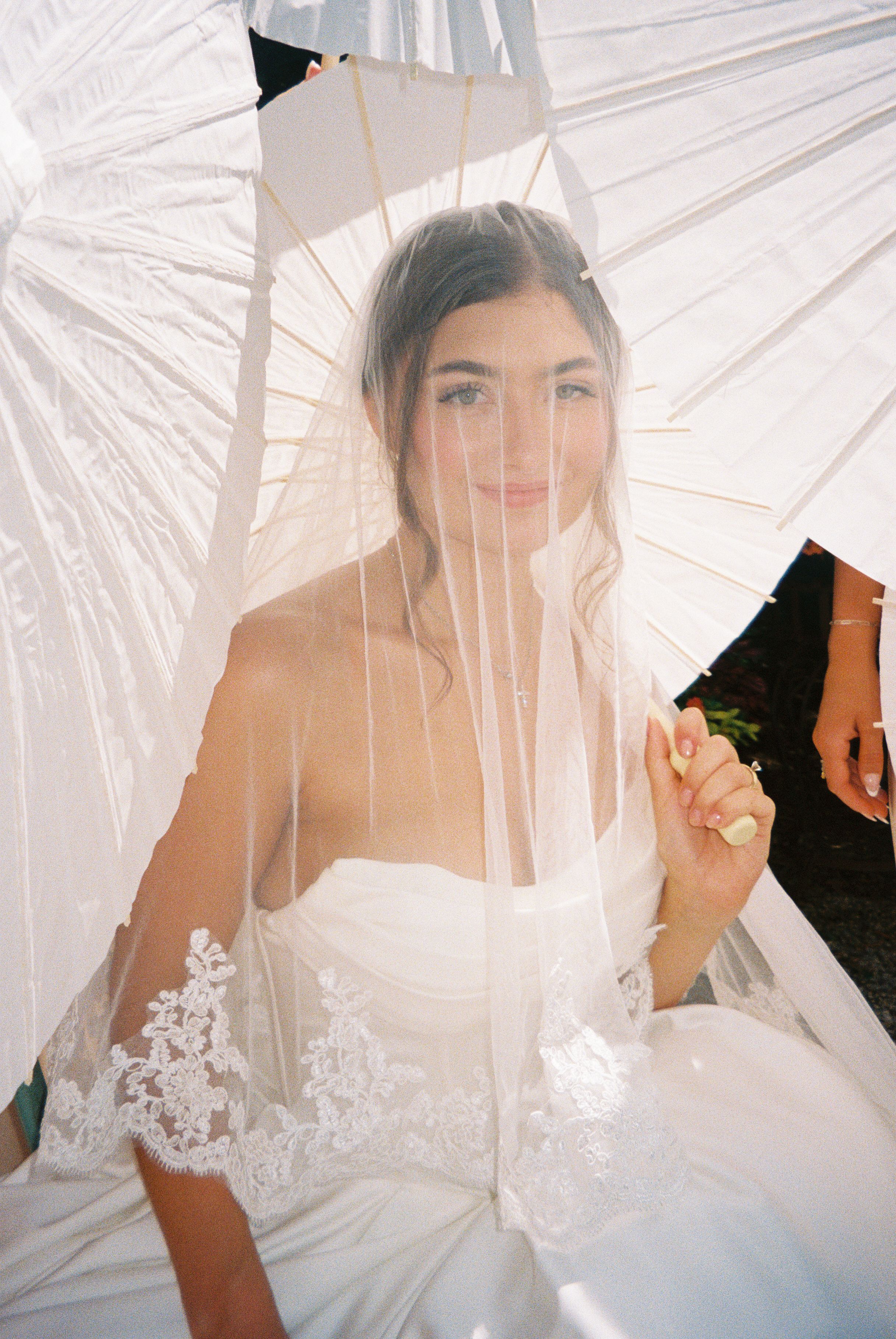 A bride in a white strapless gown poses under a delicate lace veil in soft natural lighting.