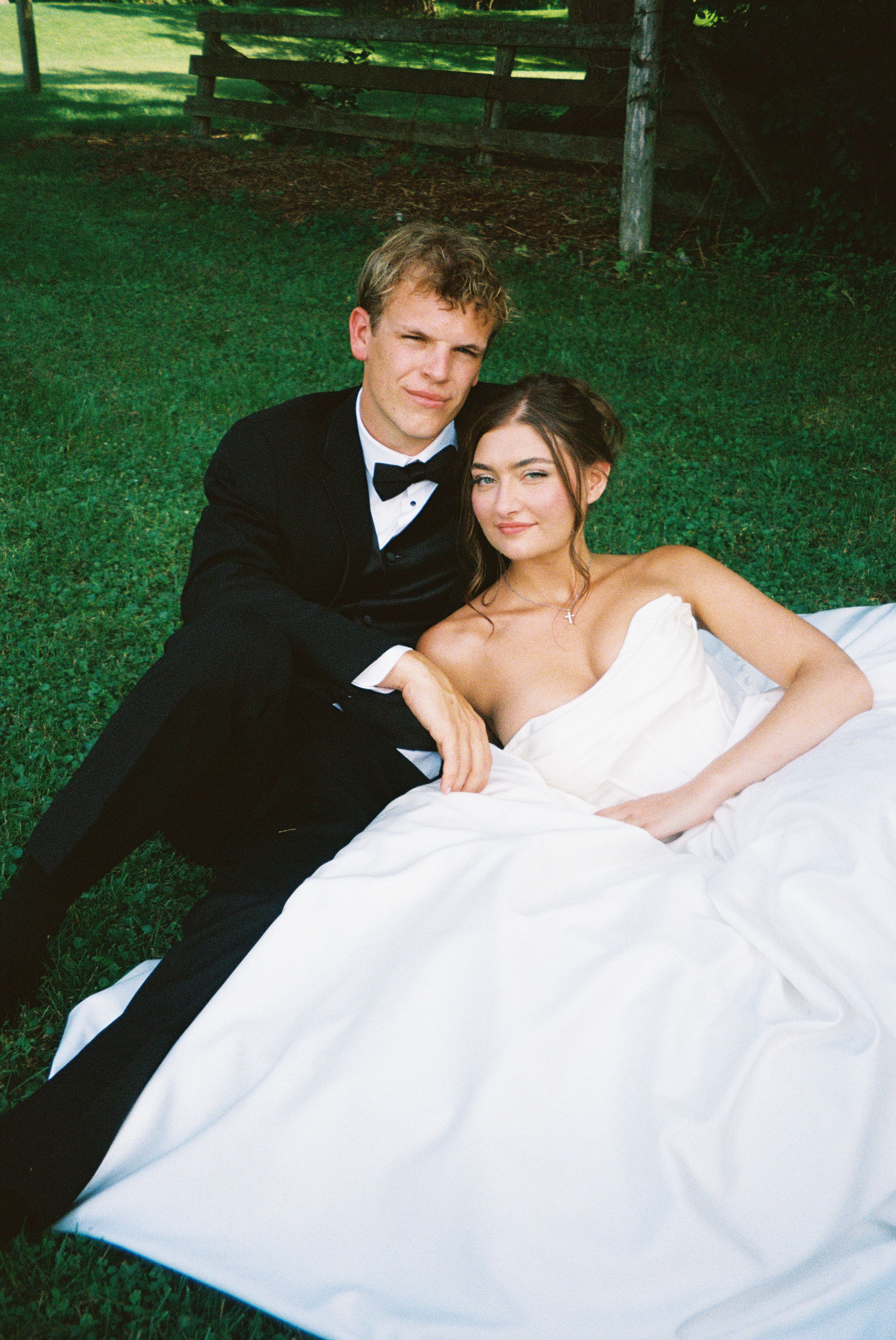 Wedding couple relaxes on grass in formal attire during an outdoor photo session.