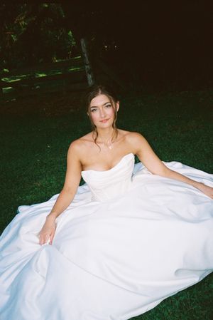 A bride in a strapless white gown sits gracefully on grass during an outdoor wedding photoshoot.