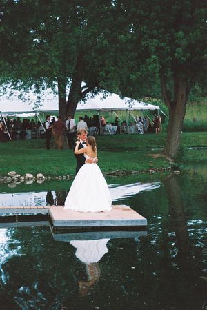 A couple shares their first dance on a dock over a reflective pond at an outdoor wedding reception.