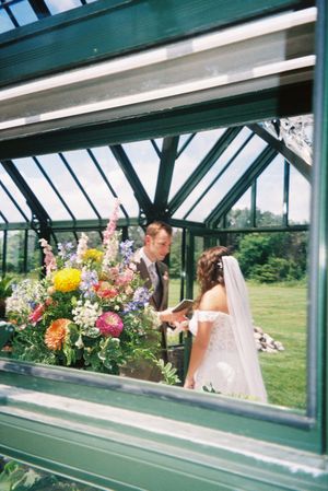 Wedding party members look through greenhouse windows surrounded by colorful floral arrangements.