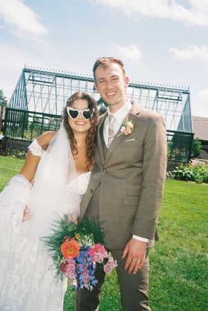 A wedding couple poses in front of a greenhouse on a sunny day with the bride holding a colorful bouquet.