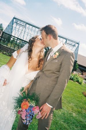 A couple shares a romantic moment in their wedding attire on a grassy lawn with a greenhouse in the background.