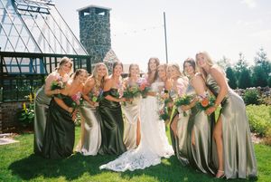 A bridal party in sage green dresses poses together in a garden setting with a stone chimney tower in the background.