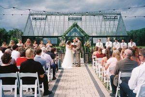 A wedding ceremony takes place under a glass greenhouse structure with guests seated in white chairs on a stone patio.