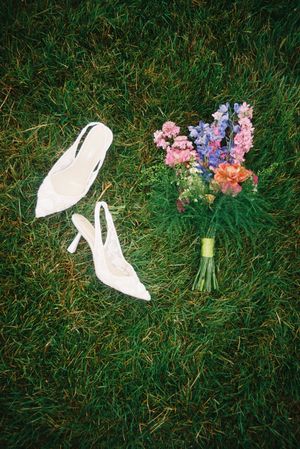 A colorful bridal bouquet with pink and blue flowers lies on green grass next to elegant white wedding shoes.
