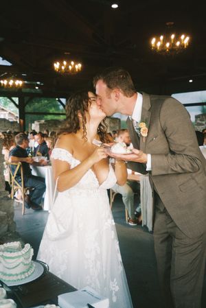 A newlywed couple shares their first bite of wedding cake during their reception under rustic chandeliers.