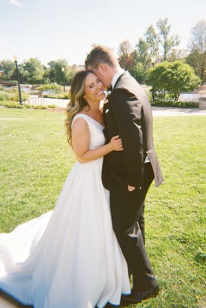 A couple in wedding attire embrace lovingly in a sunny outdoor field with trees in the background.