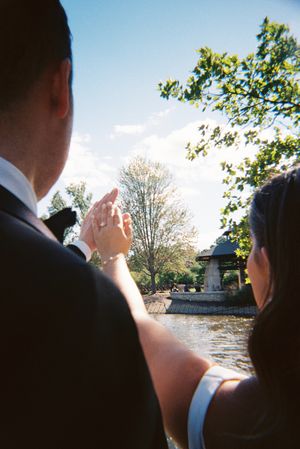 People in formal attire looking out over a water feature in a garden setting on a sunny day.