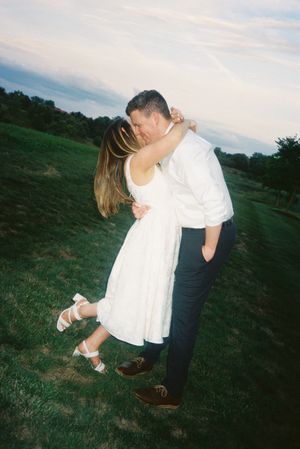 A romantic couple embraces on a grassy hillside at sunset, with the woman wearing a white dress and silver sandals.