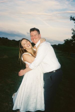 A couple in wedding attire embraces and laughs together outdoors at dusk with trees in the background.