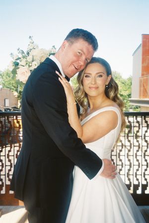 A romantic wedding portrait of a couple embracing on a balcony with white floral decorations in the background.