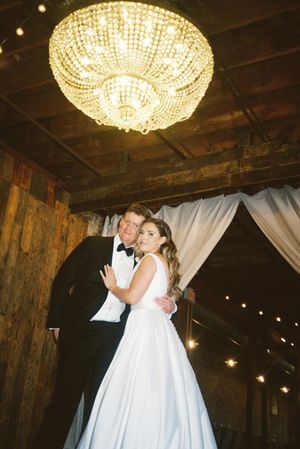 A romantic wedding moment under a crystal chandelier in a rustic wooden venue with draped white fabric and twinkling lights.