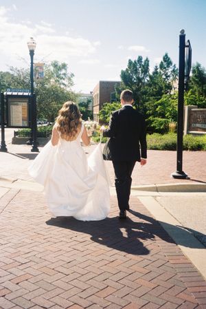 A wedding couple walking along a brick path on a sunny day with lamp posts and trees in the background.