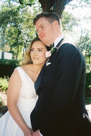 A romantic outdoor wedding portrait of a couple in formal attire standing close together under trees.