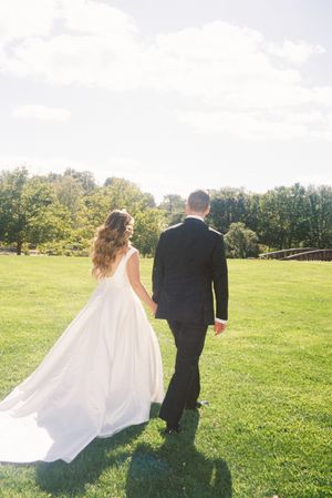 A couple walks away from the camera through a sunlit green field on their wedding day.