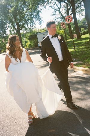 A couple in wedding attire walks together on a tree-lined path, with the bride's white dress flowing in the sunlight.