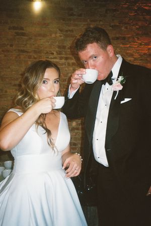 A couple in wedding attire sharing a playful moment while drinking from teacups in a rustic brick-walled setting.