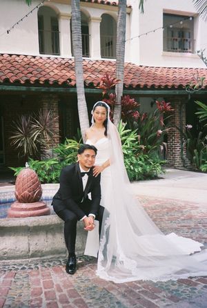 A wedding couple poses in front of a Spanish-style building with terracotta roof tiles and tropical landscaping.