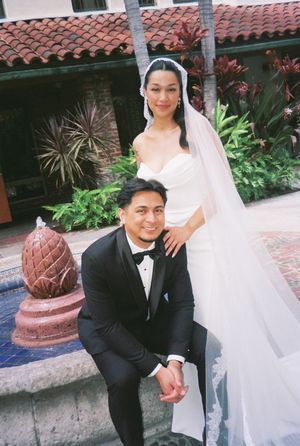 A bride in white and groom in black tuxedo pose together in a courtyard with Spanish tile roof and tropical plants.