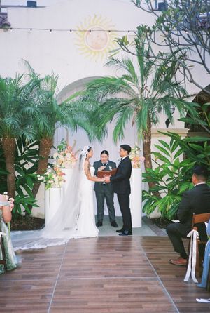 A wedding ceremony takes place under palm trees and tropical decor on a wooden deck with string lights.