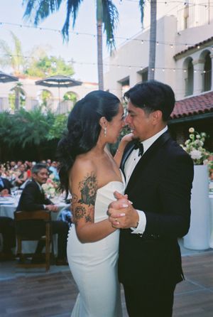 A couple shares their first dance at an outdoor wedding reception with string lights and palm trees overhead.