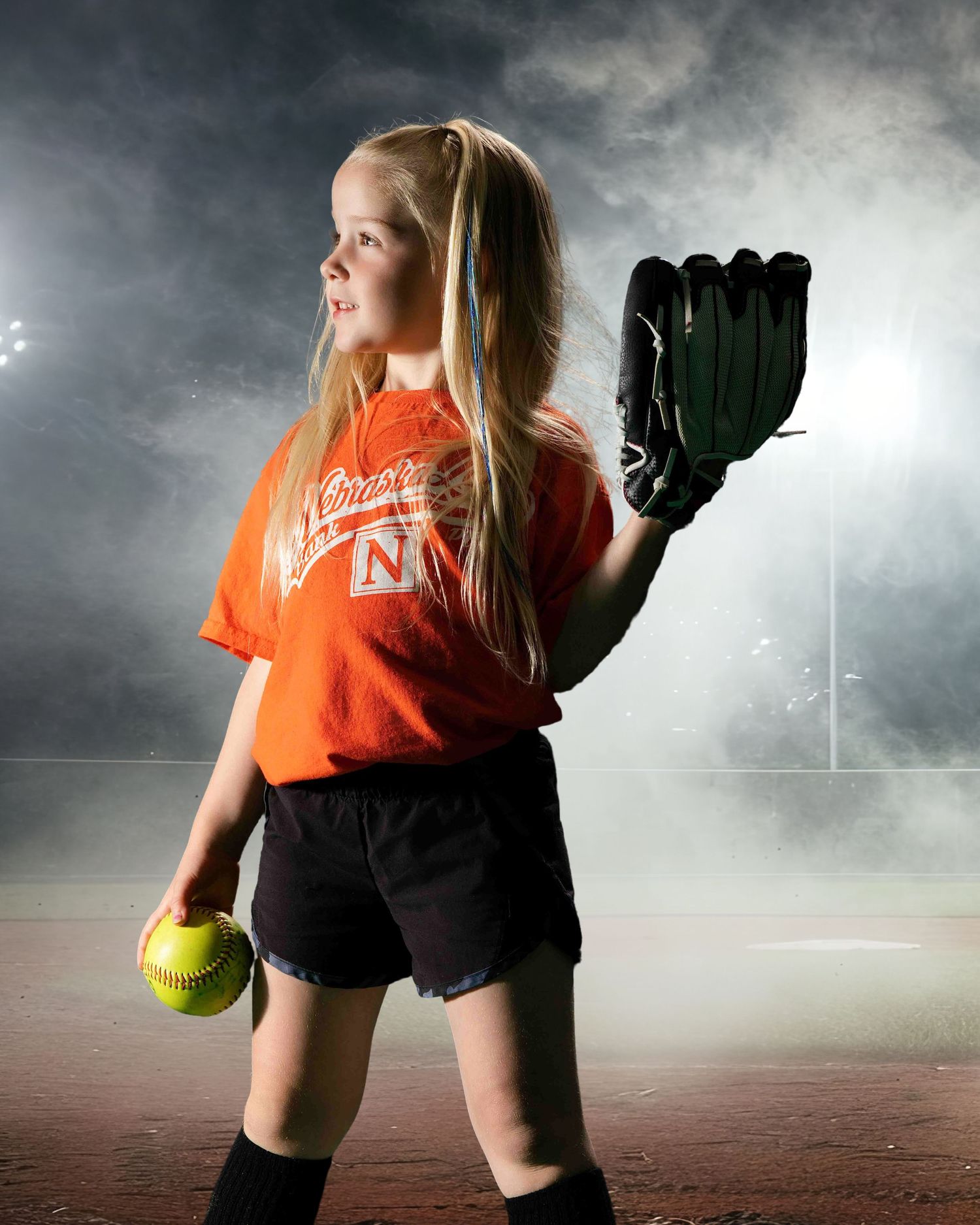 A softball player in an orange jersey stands on a misty field at dusk holding a glove and ball.