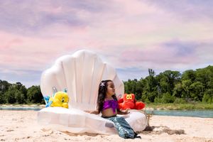White shell-shaped chair on beach with colorful props against pastel sky.