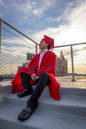 Graduate in red cap and gown reclining on rooftop during sunset.