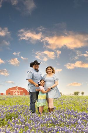 Family portrait in Texas bluebonnet field with red barn in background at sunset.