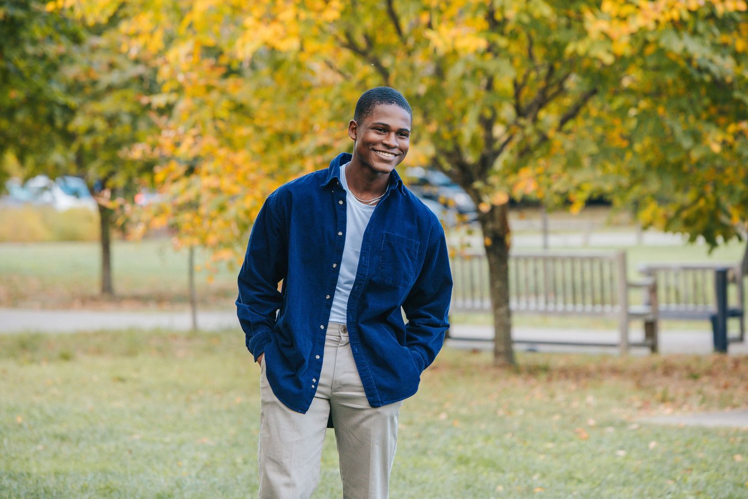 Person wearing a navy blue jacket and khaki pants stands in a park with autumn yellow trees in the background.