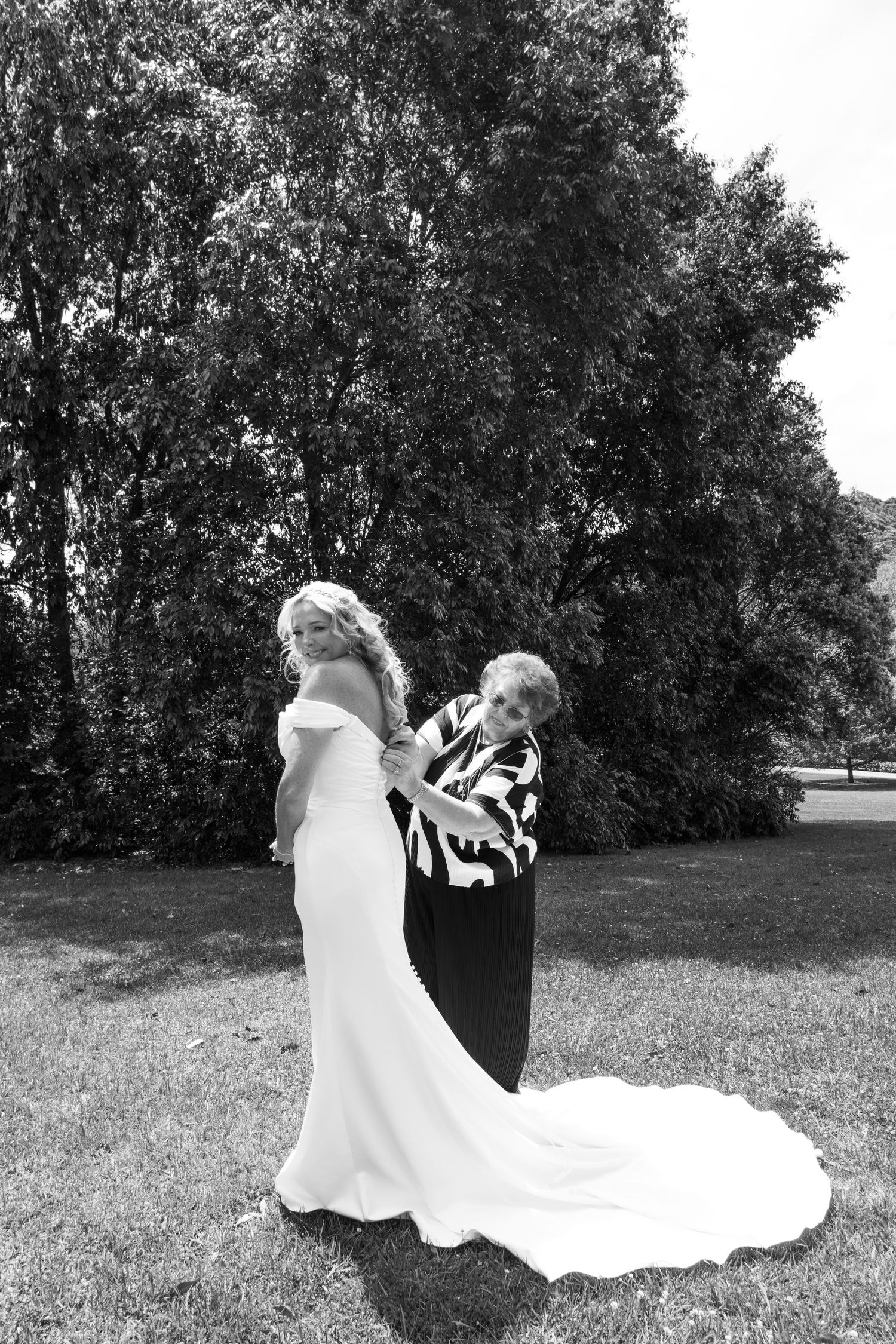 Wedding party members in black and white attire playfully pose together on a sunny grassy lawn with trees in background.