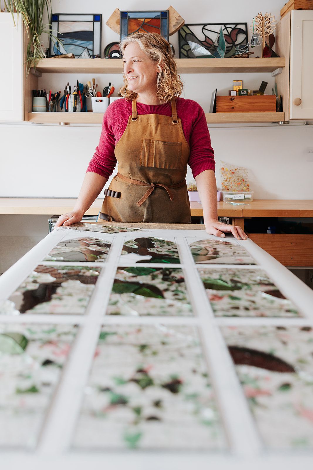 Artist wearing brown apron smiles while reviewing photo prints laid out on wooden table in bright studio space.
