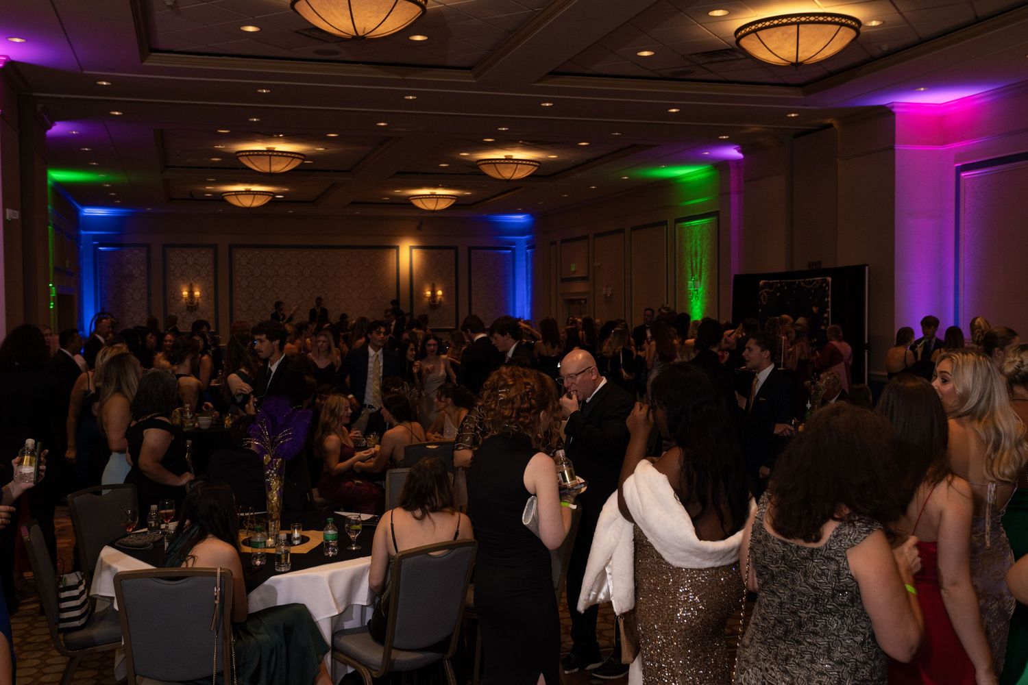 A dimly lit ballroom with colorful purple and green lighting illuminates a crowd of people during an event.